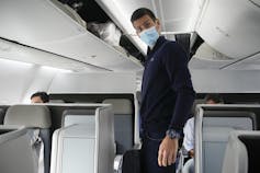 A man with dark hair in a mask stands in an airplane.