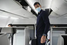 A man with dark hair in a mask stands in an airplane.