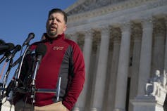 A man with a beard in a red sweater speaks into a microphone with an ornate pillared building behind him.
