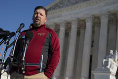 A man with a beard in a red sweater speaks into a microphone with an ornate pillared building behind him.