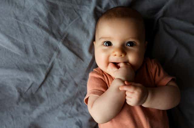 A smiling, wide-eyed infant relaxes on a bed.