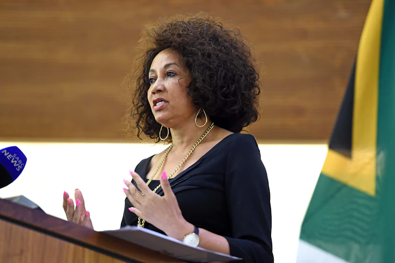 A woman with dangling earings and a flowing gold chain gestures with her open arms at chest-level as she speaks at a podium