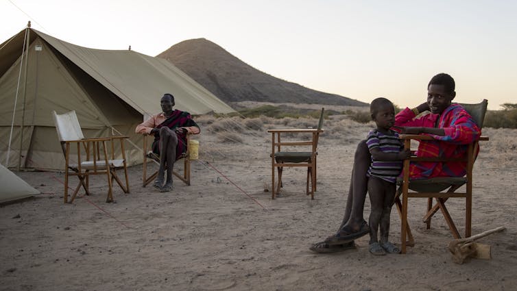 Two men and a child sitting in front of a tent with car to the right
