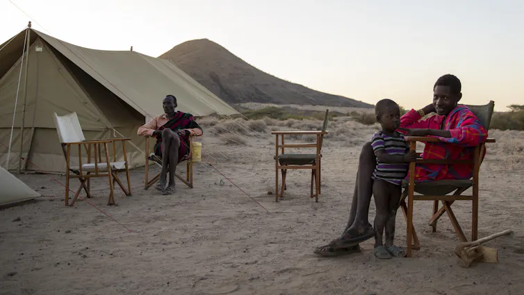 Two men and a child sitting in front of a tent with car to the right