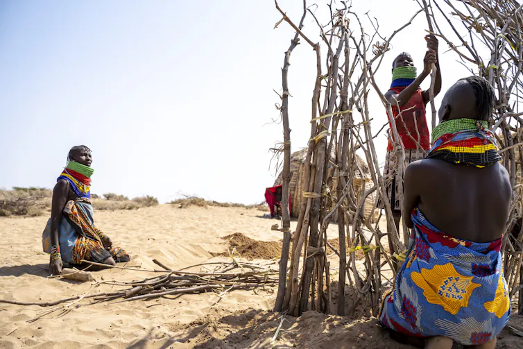 Three people tie sticks together to create a wall in a sandy landscape.
