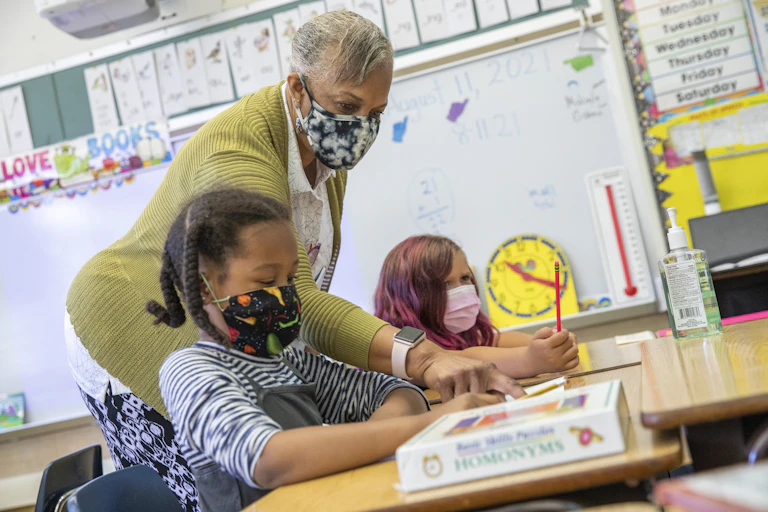 A teacher leans in to assist a student sitting at a desk.