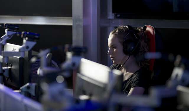 a woman wearing headphones looks at one of a row of computer monitors in a darkened room