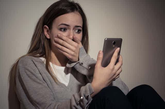 a young woman covers her mouth in shock as she looks at her phone's screen