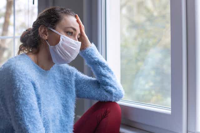 Une jeune femme en pull bleu assise à une fenêtre et portant un masque facial, l'air fatigué.