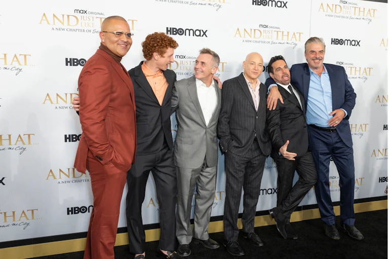 Six men stand arm in arm in front of a And Just Like That mural.