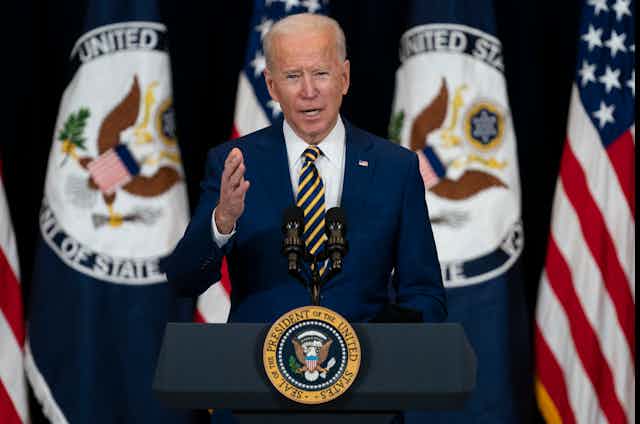 a man with white hair wearing a blue business suit stands in front of a row of flags at a podium with a seal on the front and gestures with his right hand