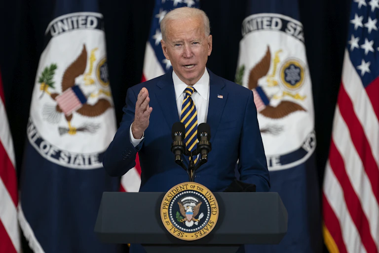 a man with white hair wearing a blue business suit stands in front of a row of flags at a podium with a seal on the front and gestures with his right hand