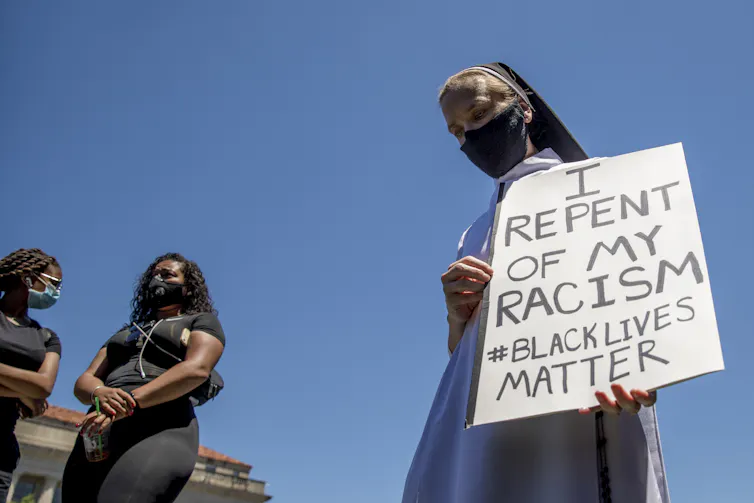 A woman holds a sign that reads I Repent of my Racism #blacklivesmatter as she prays.