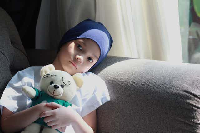 A child with cancer wearing a headscarf and holding a teddy bear