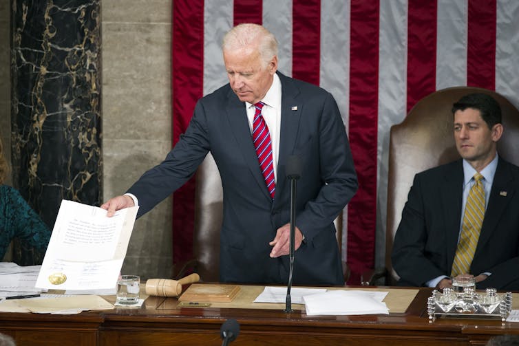 Then-Vice President Biden handing over a sheet of paper to an aide, against a backdrop of the American flag.