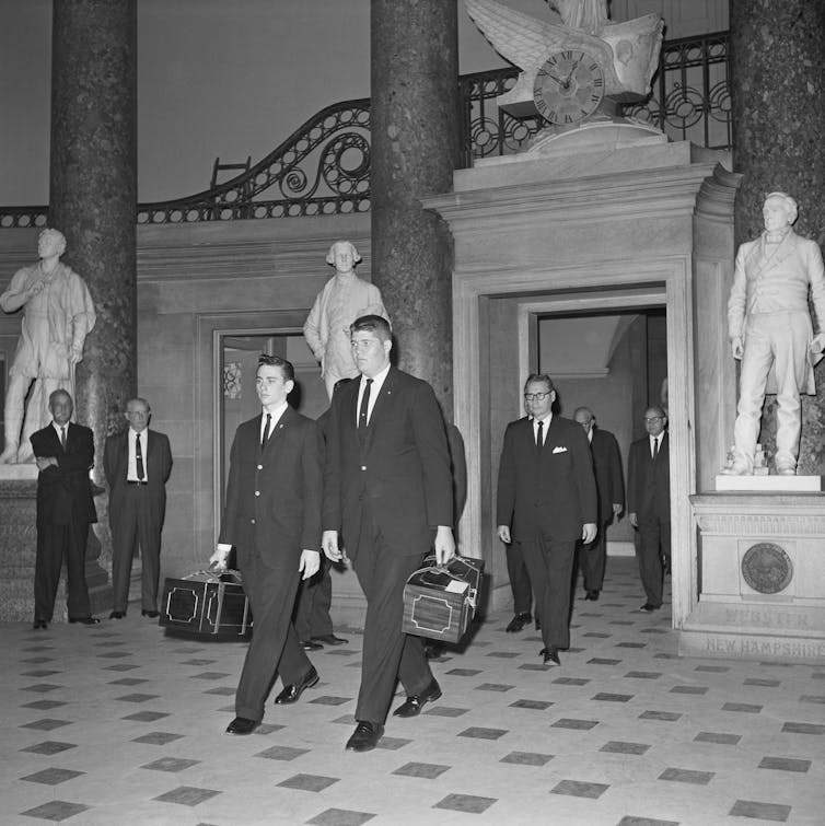 Jóvenes vestidos de traje llevan cajas de caoba a través de un salón del Capitolio de los Estados Unidos.