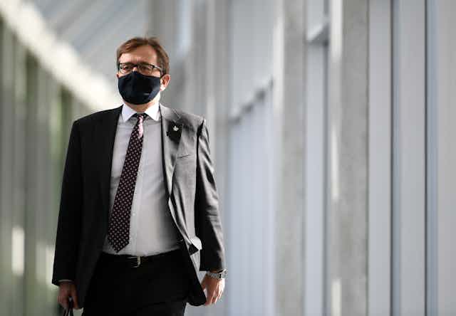 A man in a suit and a black face mask walks down a hallway.