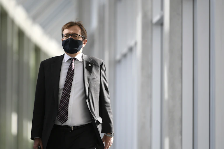 A man in a suit and a black face mask walks down a hallway.