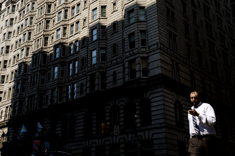 A man in a white shirt and tie looks down at his smartphone as he crosses a street with a building behind him.