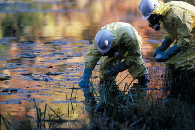 Two technicians in protective suits sample polluted river water.