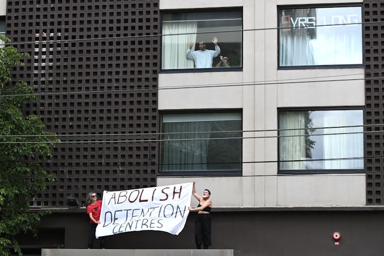 A man and a young child bang on the window from inside the Park Hotel while protesters carrying a banner reading Abolish Detention Centres stand outside.