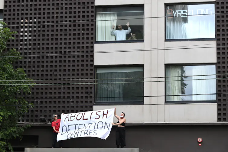 A man and a young child bang on the window from inside the Park Hotel while protesters carrying a banner reading Abolish Detention Centres stand outside.