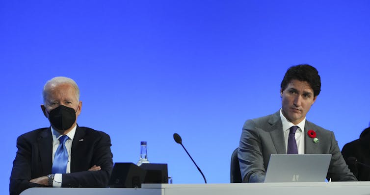 Two men sit at a table with mics and laptops in front of them.