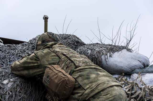 A Ukrainian soldier dressed in camouflage lying in a trench, watching Russian troops through a periscope 