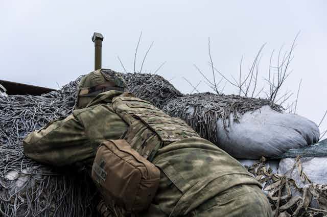 A Ukrainian soldier dressed in camouflage lying in a trench, watching Russian troops through a periscope