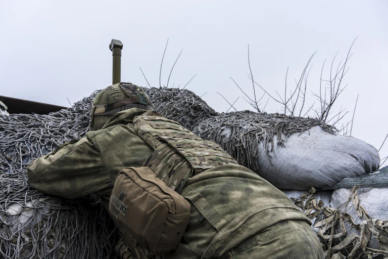 A Ukrainian soldier dressed in camouflage lying in a trench, watching Russian troops through a periscope