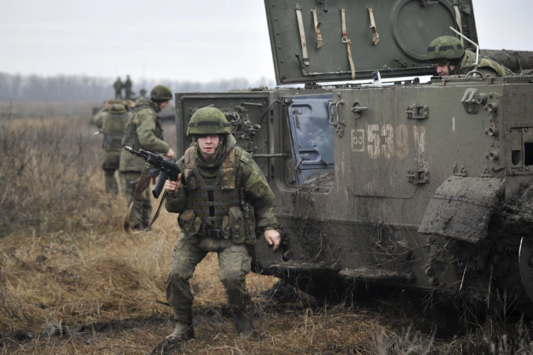 A young soldier clad in green military clothing crouches and points a gun. The soldier is standing in front of a tank