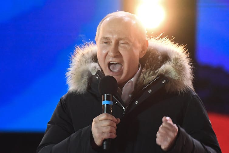 Vladimir Putin in a winter coat with a fur collar, speaking into a microphone at a rally and looking excited and happy.