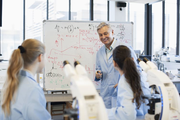 man at white board, two women facing him with microscopes in foreground