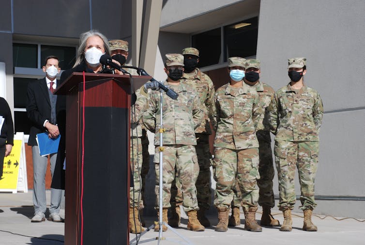 A person speaks at a lectern, with several people in camouflage uniforms behind her.