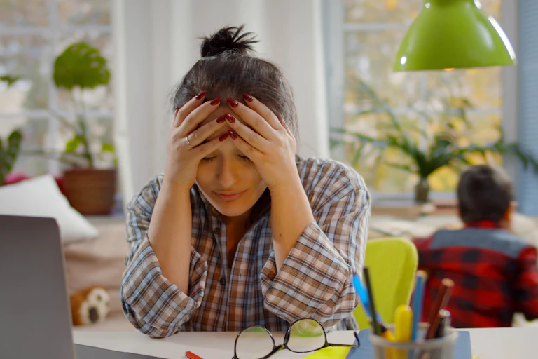 Woman with head in hands struggling to study as children play in the background