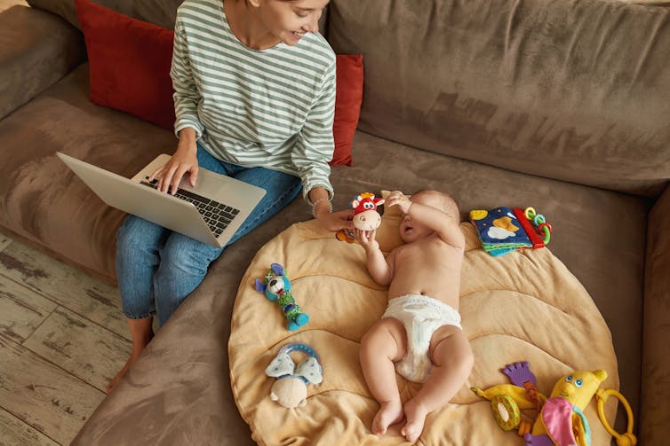 mother with laptop on her knee offers a toy to a baby in nappies next to her