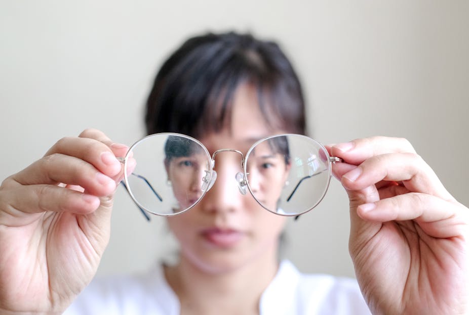 Woman holds her prescription eyeglasses in front of her and looks through the lenses.