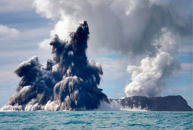 Plumes of steam, smoke and ash tower above tropical ocean water.