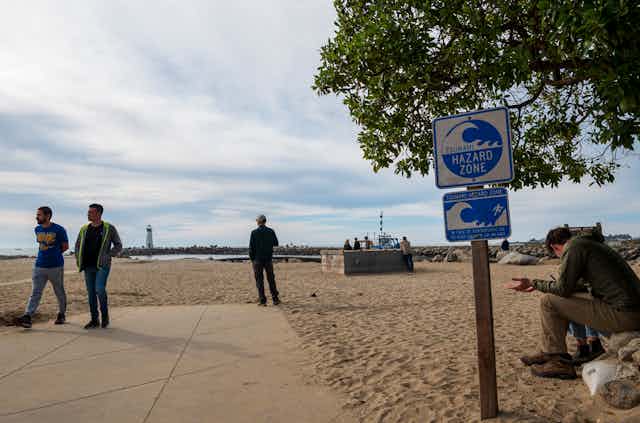 people walk on a beach with a TSUNAMI HAZARD ZONE sign