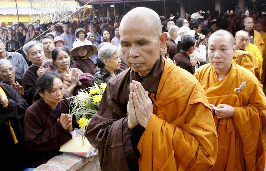 A Buddhist monk in saffron-colored robes folds his hands in prayer accompanied by worshipers and similarly clad monks.