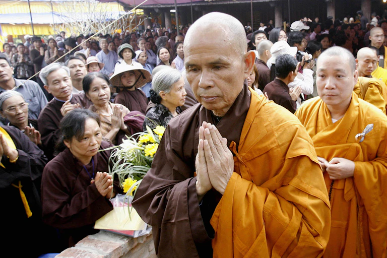 A Buddhist monk in saffron-colored robes folds his hands in prayer accompanied by worshipers and similarly clad monks.