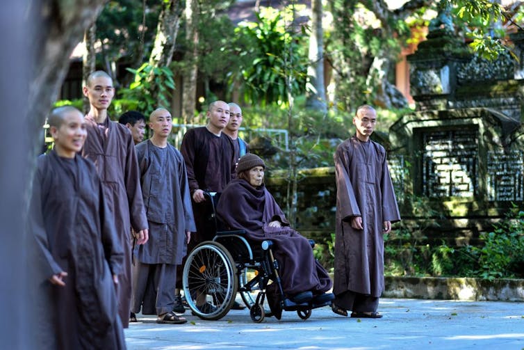 El monje budista Thich Nhat Hanh en una silla de ruedas con una túnica morada y rodeado de monjes con túnicas similares.