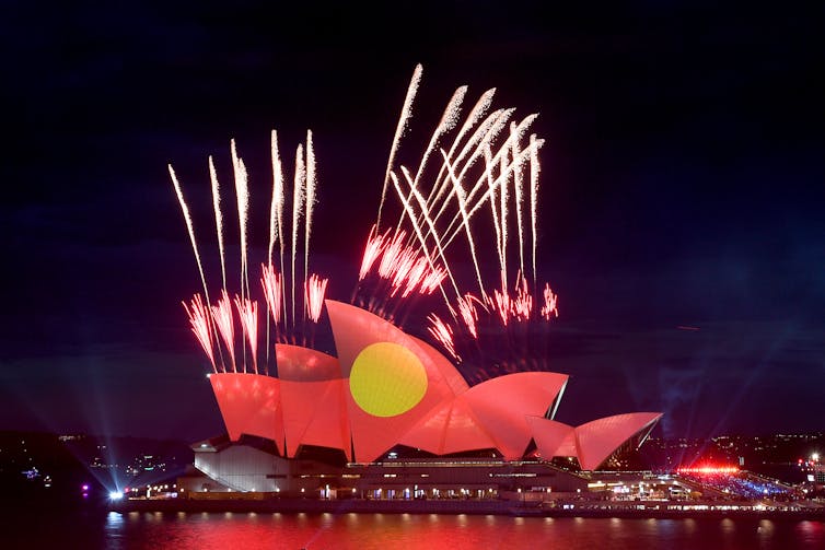 Fireworks go off over the Opera House, illuminated with the Aboriginal Flag.