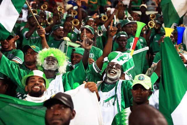Fans with painted faces and dramatic hats in the Nigerian colours, green and white, cheer and smile