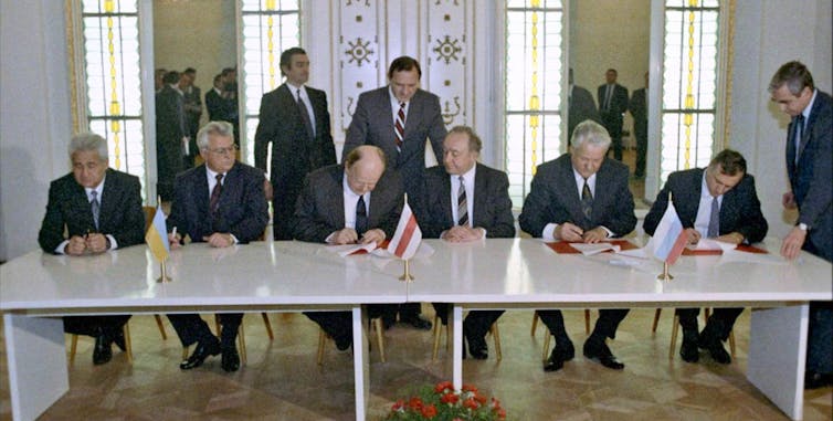 Several men in suits sit at a long table, some of them signing documents.