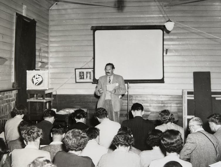 A man stands in front of a screen, talking to a crowd