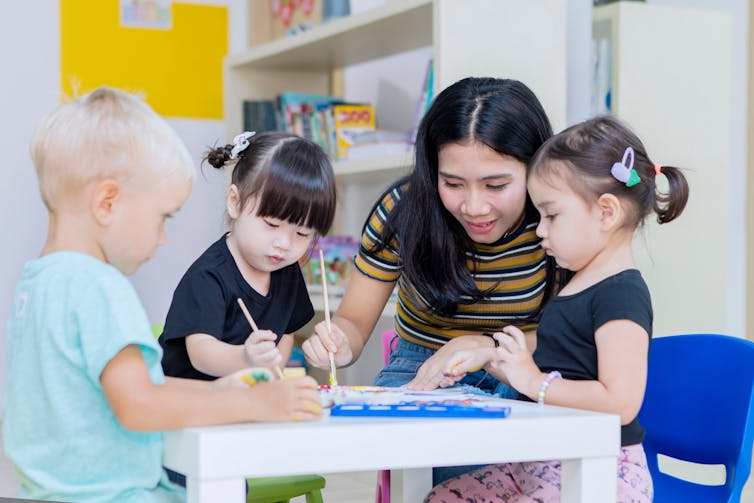 A woman is seen with children at a desk guiding painting.