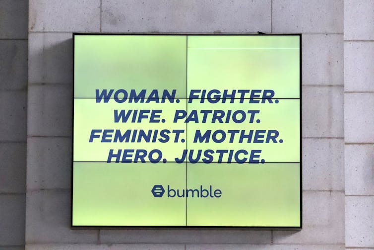 A video billboard reads 'Woman. Fighter. Wife. Patriot. Feminist. Mother. Hero. Justice.'