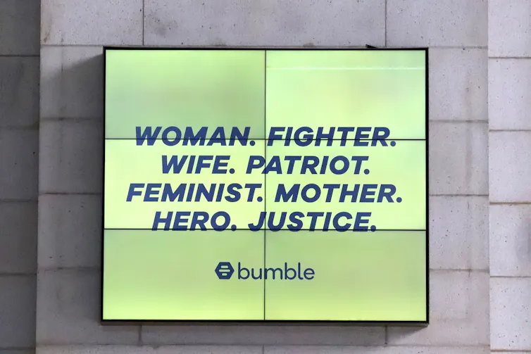 A video billboard reads 'Woman. Fighter. Wife. Patriot. Feminist. Mother. Hero. Justice.'
