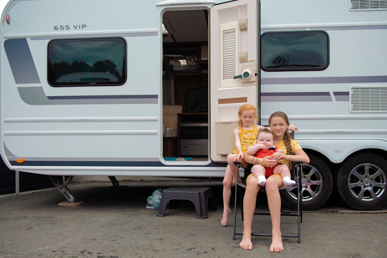 Three young Traveller children seated on a folding chair outside of a caravan